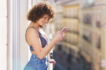 Side view of positive young female in bra and jeans with curly hair smiling while browsing smartphone on sunny balcony. Cheerful woman using smartphone on balcony