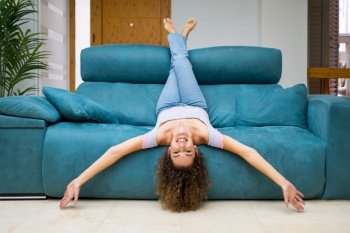 Smiling young barefoot female with curly hair lying upside down on couch and looking at camera while having fun at home. Happy woman woman lying upside down on couch at home
