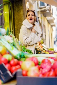 Low angle of cheerful young woman in warm clothes, smiling and looking at camera while standing near stall with bunch of fresh ripe fruits and vegetables in daylight. Gleeful young woman in cozy coat standing by fruits in shop