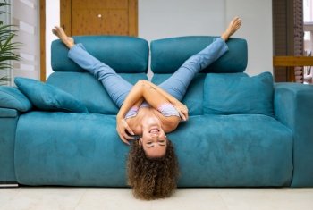 Happy young female in jeans and top with curly hair lying upside down on sofa in living room and having fun at home while enjoying. Cheerful woman lying upside down on sofa at home