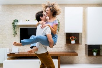 Side view of cheerful young couple in casual clothes smiling and hugging while having fun at home. Happy young couple embracing while having fun at home