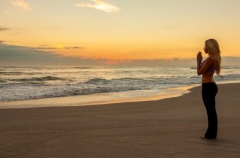 Young fit blonde woman alone practicing yoga on a beach at sunrise or sunset