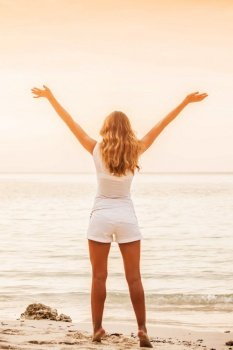 Beautiful young woman standing on sandy beach against orange sky at sunset. Summer travel. Happy slim girl in white with raised up arms on the seashore. Woman on beach at sunset