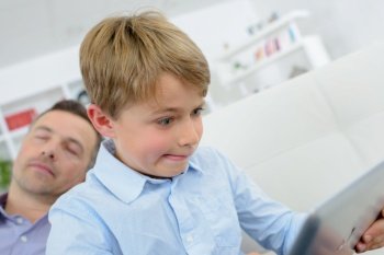 Child making funny expression, looking at laptop while father asleep