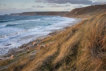 Beautiful landscape of Sennen Cove in Cornwall during sunset viewed from grassy sand dunes with moody sky and long exposure sea motion
