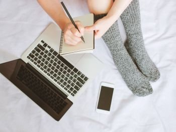 Top view of woman legs in socks and writing on notebook over laptop and smartphone on cozy bed, lifestyle concept, autumn and winter season