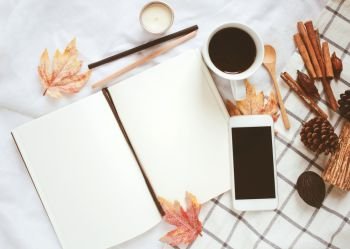 Autumn lifestyle concept, blank notebook, smartphone and coffee with autumn leaves ornaments on white bed sheet background