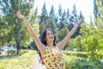 Beautiful long-haired brunette in a light silk dress with polka dots with his hands up she rejoices in the sun against the backdrop of summer nature