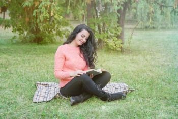 Beautiful long-haired brunette girl in a warm pink sweater sits in the park and reads a book