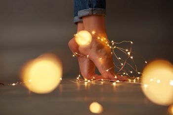 Christmas garlands on the floor are decorated with female feet around a dark background with a blur of yellow lights. Copy space. Feet of a woman decorated with yellow garlands around a dark background