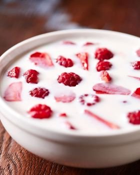 Delicious fresh food with white milk and red raspberries, strawberriees in a white bowl on a wooden background.. White bowl with helthy breakfast with fresh ripe berries and yoghurt on a wooden table.
