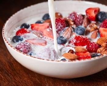 Milk is pouring into homemade natural breakfast with fresh organic ingredients - strawberry, raspberry, blueberry, granola, nuts in a white bowl on a wooden background. Healthy vegetarian eating.. Preparation of dietary natural breakfast with fresh organic ingredients - berries, granola, nuts, honey and pouring milk in a white bowl on a wooden table.