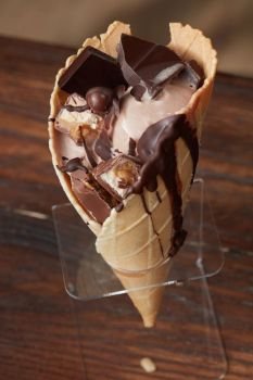 Waffle cone with melted ice cream chocolate syrup and pieces of chocolate in a stand, on a wooden dark background. Ice cream in waffle cone