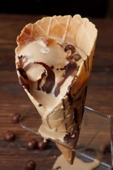 Melting ice cream with chocolate topping in a waffle cone on a stand on a wooden brown background. Ice cream in waffle cone