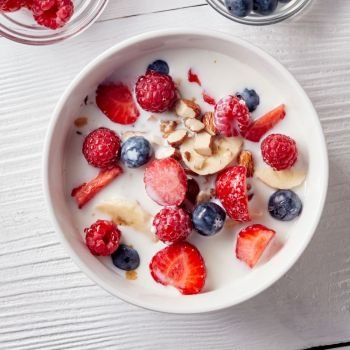 Close-up of a white ceramic bowl on a white table with natural organic ingredients for natural healthy breakfast. Concept of natural clean eating. Flat lay.. Cereal muesli and sliced bananas, strawberries, berries, chopped almonds and walnuts with milk in a white ceramic bowl on white table. Top view.