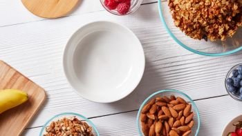 Empty white ceramic bowl on a white table with natural ingredients for preparing delicious healthy organic breakfast. Concept of healthy eating.. Ingredients for preparing healthy breakfast - berries, cereal flakes, muesli, banana, honey, almonds, walnuts on white. Top view.