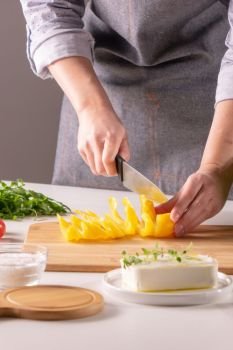 Fresh cheese with herbs, salt, rucola on white kitchen table. Female hands cut the pepper into pieces to make a salad. Step-by-step recipe. Pieces of pepper on a wooden board, female hands cut vegetable for salad on a white kitchen table