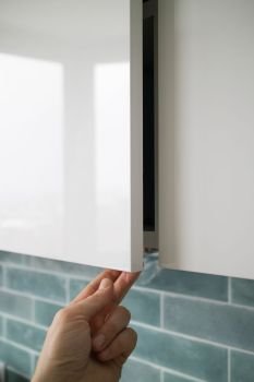 The closeup man hand opening or closing kitchen cabinet door on a background of blue kitchen interior. Modern kitchen furniture without handles with an additional small lower for opening the door. man hand opening or closing kitchen cabinet door