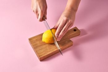 Citrus fruits on cutting board. Female hands cutting a yellow ripe lemon on half on a wooden board on a pink background. Concept vegetarian food.. A girls’ hands cut a yellow ripe lemon in half on a wooden board on a pink background.