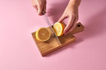 Citrus fruits on brown board cutting by female hands. Top view of citrus fruit - slices of of yellow organic lemon on a wooden board on a pink. Healthy food concept.. Slices of yellow lemon on a wooden board on a green. Female hands cutting a ripe lemon for preparing homemade fresh cocktail on pink background..