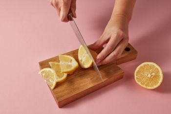 Woman cutting organic yellow ripe lemon with steel knife on pink for preparation detox drink for dietary healthy food. Concept vegetarian food.. Citrus fruit natural lemon on cutting board. Female hands cutting a yellow ripe lemon on half on a wooden board on a pink background.