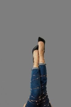 A girls legs tied with a Christmas garland in shoes and jeans on a gray background with copy space.. Legs up a young woman in blue pants and shoes with a New Year’s glowing ribbon on a gray.