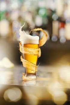 A cold glass of beer with beer foam and splash around on a wooden bar counter and blurred background with many bokeh. A cold glass of beer and a splash around on a blurred bar counter background