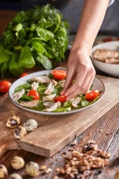 The hand of the girl puts in a plate with spinach and meat pieces of juicy tomato on an old wooden table. Step by Step cooking healthy salad.. Woman’s hand puts a piece of tomato in a plate with spinach and meat on a wooden table. Salad preparation.