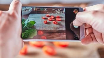 Woman’s hands are shooting on a smartphone photo of sliced tomatoes on a wooden board. Home made food photo for social networks.. Sliced tomato on a wooden board. Woman’s hands with a smartphone take off cooking salad. Food blogger concept