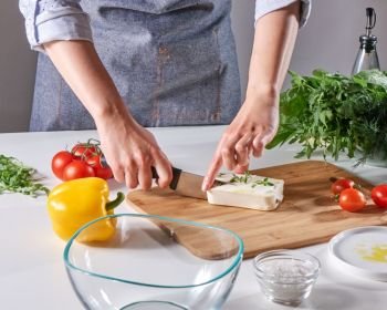 Hands of a cook girl are cutting organic cheese with sprouts on a wooden board on a table with an empty bowl and vegetables. Step by step preparation. Woman’s hands cut the cheese with the sprouts on the wooden board on the kitchen table with different vegetables. Step by step cooking healthy salad