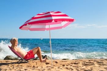 Senior man sun bathing at the beach under parasol with waves, sea and sand