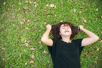 Young Asian woman laying on the green grass listening to music in the park with a chill emotion. Young woman relaxing on the grass with her music playlist. Outdoor activity in the park concept.