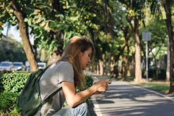 Portrait of attractive happy asian woman holding smartphone while sitting on roadside at the city street. Woman is sitting on the street near park. Summer holidays, road trip, vacation, travel concept