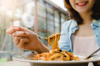 Beautiful happy Asian woman eating a plate of Italian seafood spaghetti at restaurant or cafe while smiling and looking at food.