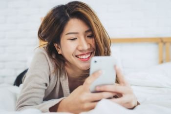 Happy Asian women are using smart phone on the bed in morning. Asian woman in bed checking social apps with smartphone. Smiling woman surfing net with cellphone at home. Mobile addict concept. 