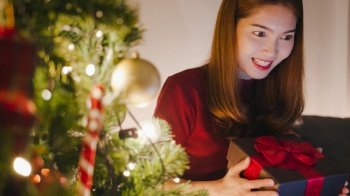 Young Asia female using tablet video call talking with couple with X’Mas present box, Christmas tree decorated with ornament in living room at home. Christmas night and New Year holiday festival.