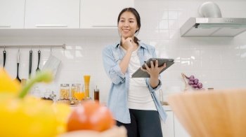 Happy Asian woman using tablet for looking recipe while making food in the kitchen, female use organic vegetable for healthy food at home. Lifestyle women making food concept.