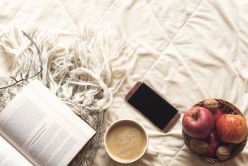 Coziness concept with an open book on a wool shawl, a basket with apples and nuts, a phone and a cup of hot cocoa on a fluffy, fleece blanket.