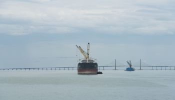 A landscape image of a cargo ship sailing past the Penang Bridge. A landscape image of a cargo ship sailing past the Penang Bridge.. A landscape image of a cargo ship sailing past the Penang Bridge.