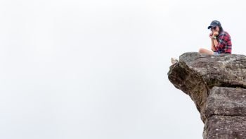 Asian woman sitting on a rock near a cliff on a white fog background is pretending to be thrilled, 16:9 widescreen. Woman on the cliff