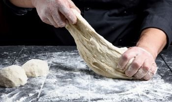 chef in black uniform kneads white wheat flour dough on a black wooden table