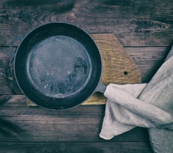black round frying pan with wooden handle and gray linen kitchen napkin on a brown wooden table, top view, vintage toning