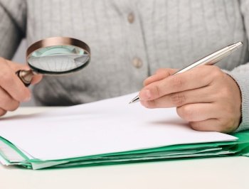 a woman sits at a white table and holds a metal pen over a pile of papers, in the other hand a magnifying glass. Finding mistakes, analyzing the budget
