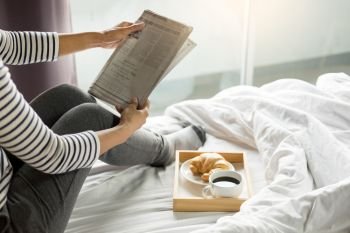 Woman reading book or newspaper and drinking coffee breakfast on bed during the morning.