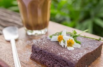 Chocolate Brownie Cake and Latte Coffee and Daisy Flower on Chopping Board or Cutting Board on Wood Table on Green Tree Background Right Frame.