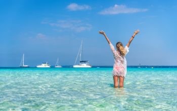 Beautiful young woman in white lace dress with raised up arms in transparent sea at sunny day in summer. Tropical landscape. Sexy back of slim girl, clear water, yachts, sky. Tanned woman on the beach