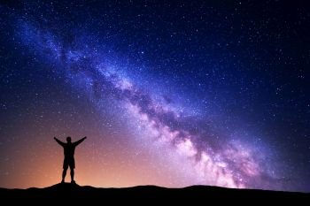 Purple Milky Way with silhouette of a standing man with raised up arms on the hill. Night landscape with beautiful galaxy. Universe. Night starry sky with Milky Way and yellow light