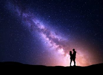 Milky Way with silhouette of people. Landscape with night starry sky. Standing man and woman on the mountain with yellow light. Hugging couple against purple milky way. Beautiful galaxy. Universe
