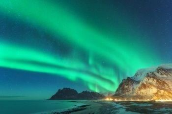 Northern lights in Lofoten islands, Norway. Green Aurora borealis. Starry sky with polar lights. Night winter landscape with aurora, sea with sky reflection, rocks, beach and snowy mountains. Travel