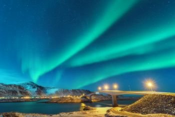 Northern lights above bridge with illumination in Lofoten islands, Norway. Aurora borealis. Starry sky with polar lights. Night winter landscape with aurora, road, village and snowy mountains. Travel